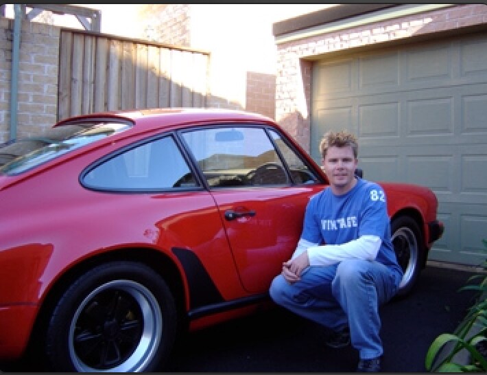 A young Lee Powell crouched beside his red Porsche 911 SC